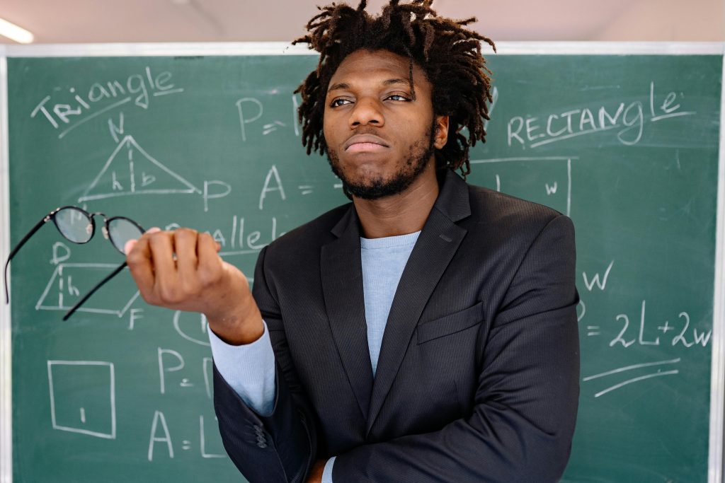 Educator posing with eyeglasses in hand, chalkboard filled with geometric formulas.